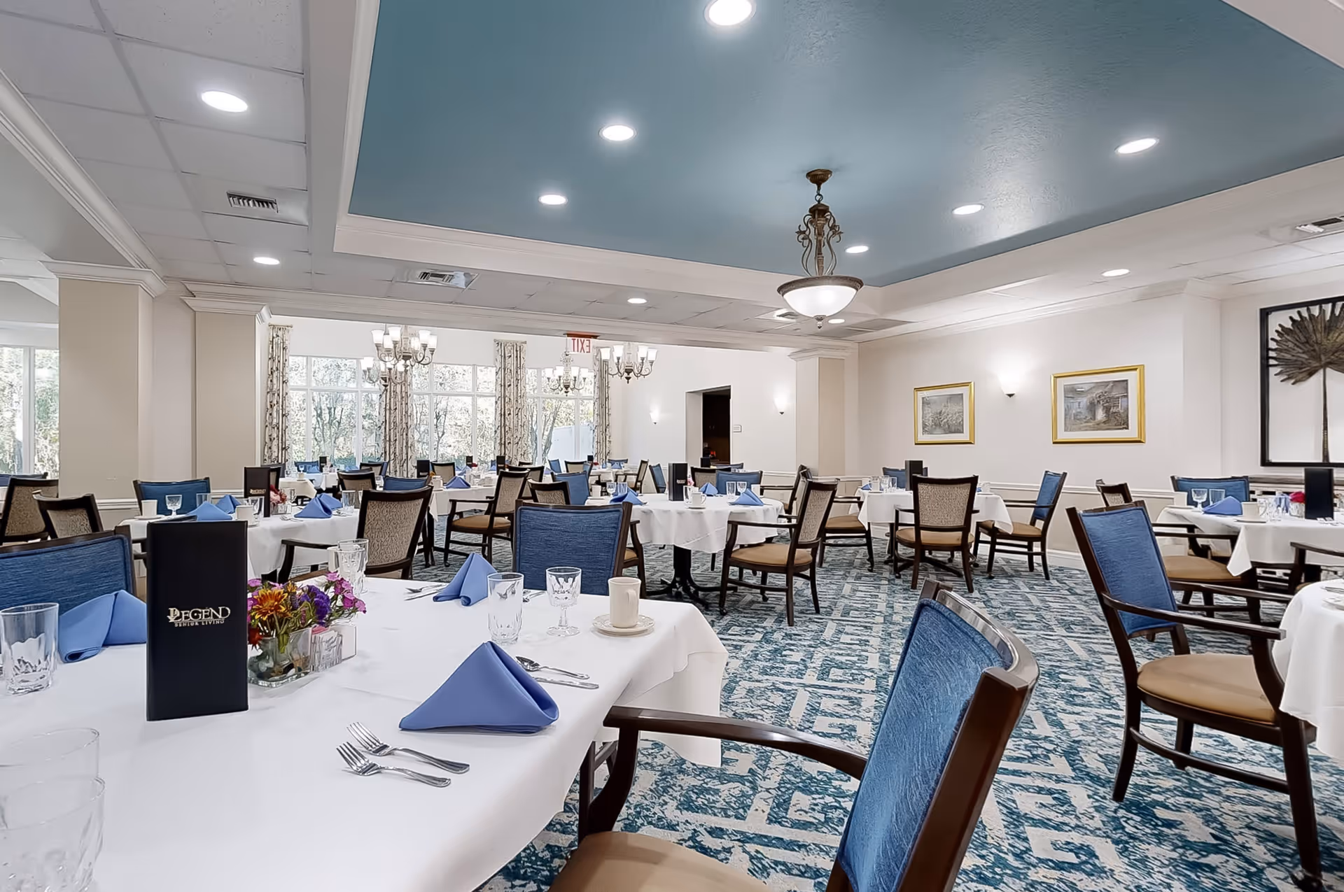 A spacious and elegant dining room with multiple tables covered in white tablecloths, each set with blue folded napkins, glassware, and silverware. The room features blue upholstered chairs, a patterned blue and white carpet, large windows with curtains allowing natural light, and decorative light fixtures on the ceiling and walls. Framed artwork is displayed on the walls.