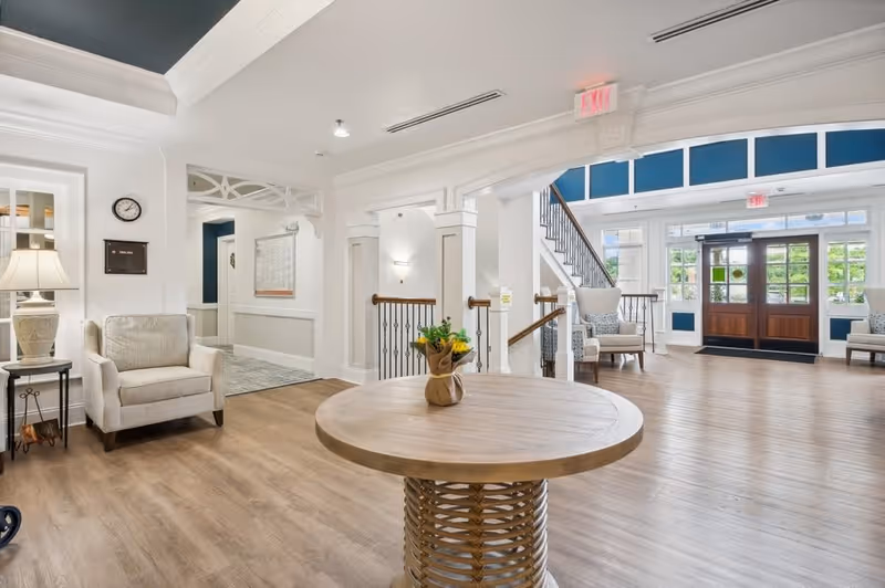 Bright and spacious senior living facility lobby with wooden flooring, a round wooden table with a small flower arrangement in the center, comfortable armchairs, and large windows letting in natural light. The entrance door is visible in the background along with a staircase and exit signs.