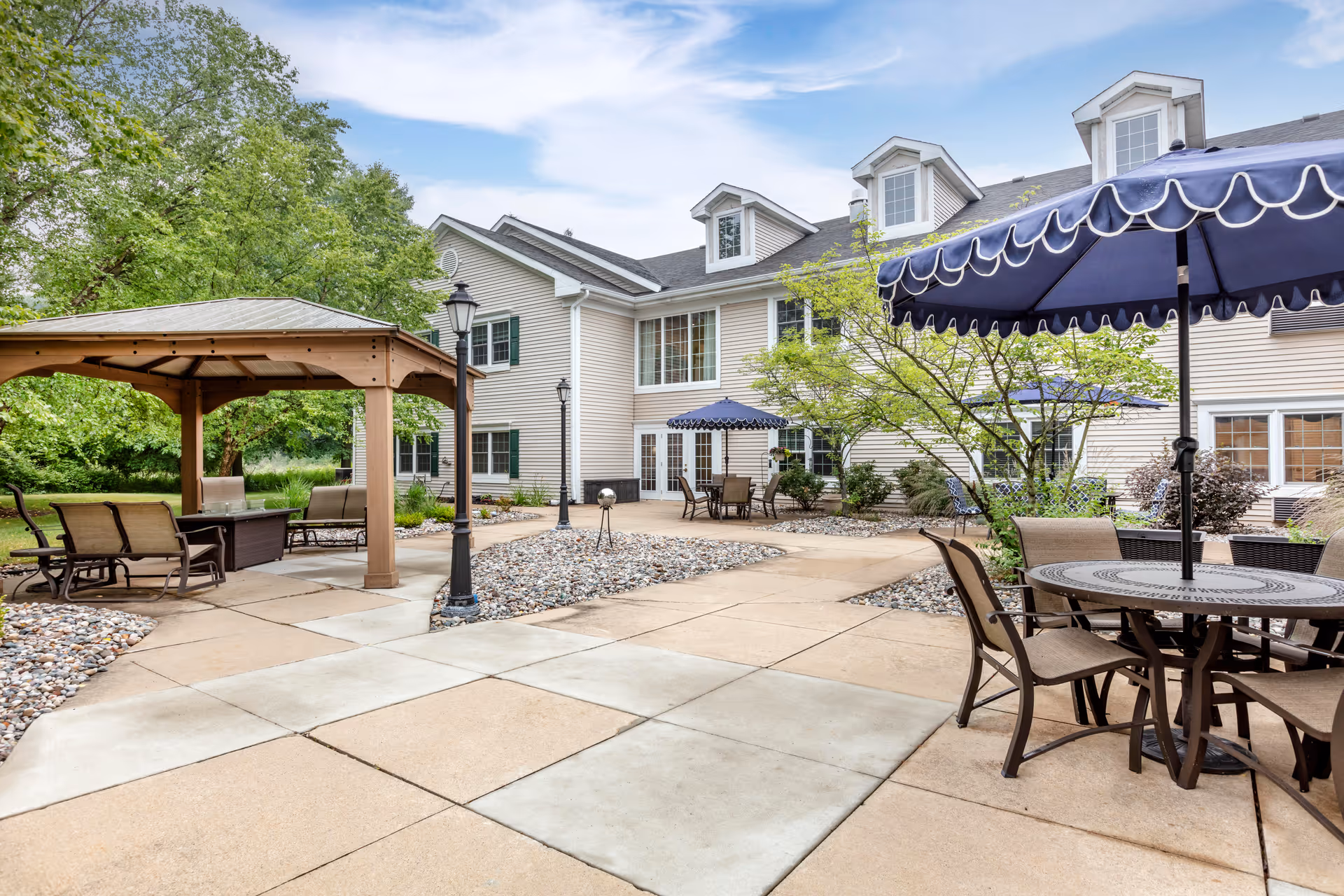 Outdoor patio area at Brookdale Meridian - Assisted Living with multiple seating arrangements including tables with blue umbrellas and a wooden gazebo. The patio is surrounded by a two-story building with white siding and several windows. There are trees and landscaping with rocks around the patio.
