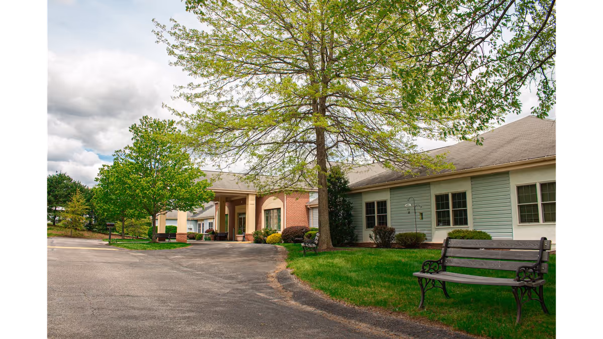 Front entrance of a single-story senior living facility with a covered portico, trees, benches, and a driveway.