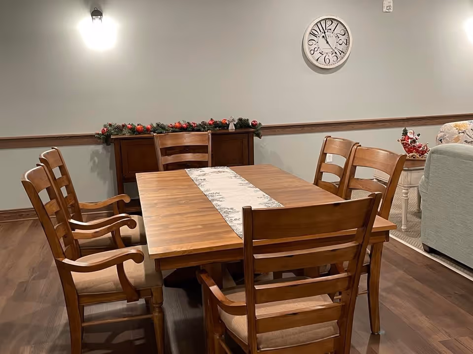 A wooden dining table with six chairs in a communal dining area decorated with garland and a wall clock.