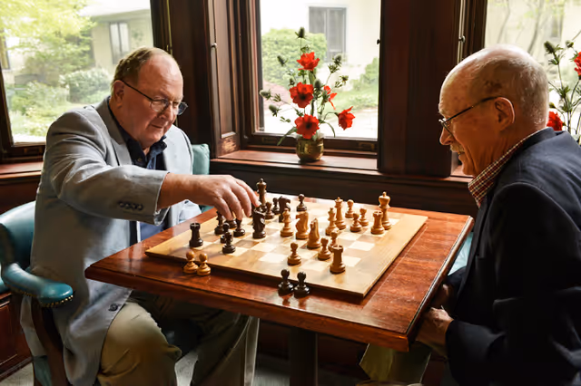 Two elderly men sitting at a wooden table playing chess in a room with large windows and a vase of red flowers on the windowsill.