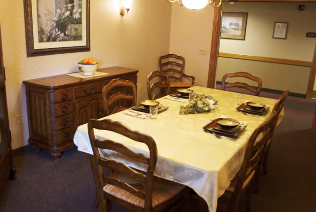 Wooden dining table set for six in a warmly lit room with matching chairs, a sideboard, and framed artwork.