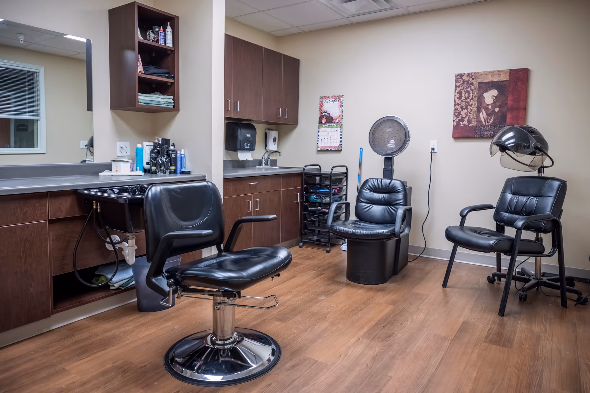 Salon-style interior room with styling chairs, a shampoo sink, hooded hair dryer, and cabinetry.