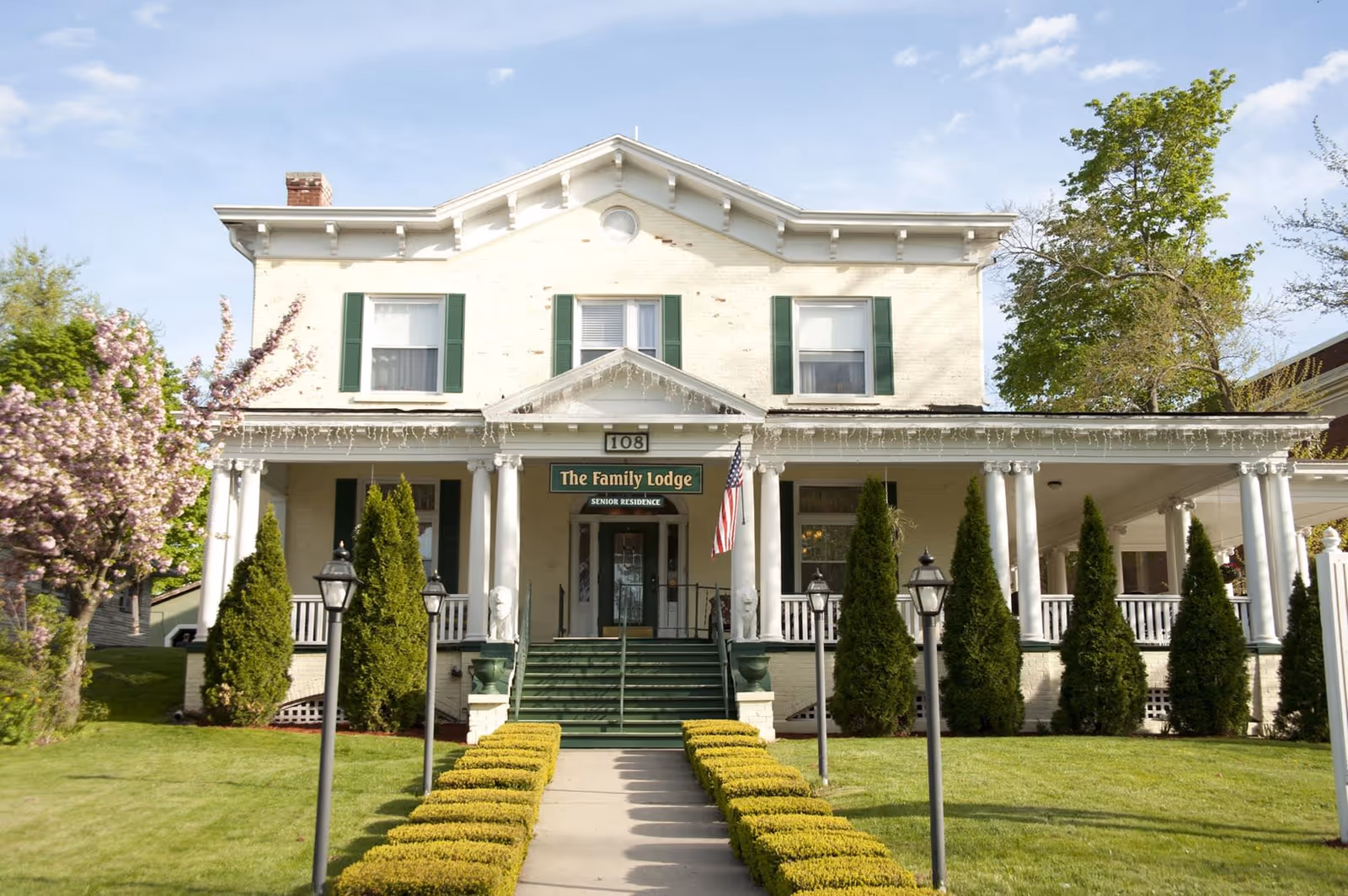 Front exterior view of a two-story senior residence building named The Family Lodge with white brick walls, green shutters, a green staircase, and a porch with white columns. The building is surrounded by neatly trimmed bushes, trees, and a lawn under a blue sky.