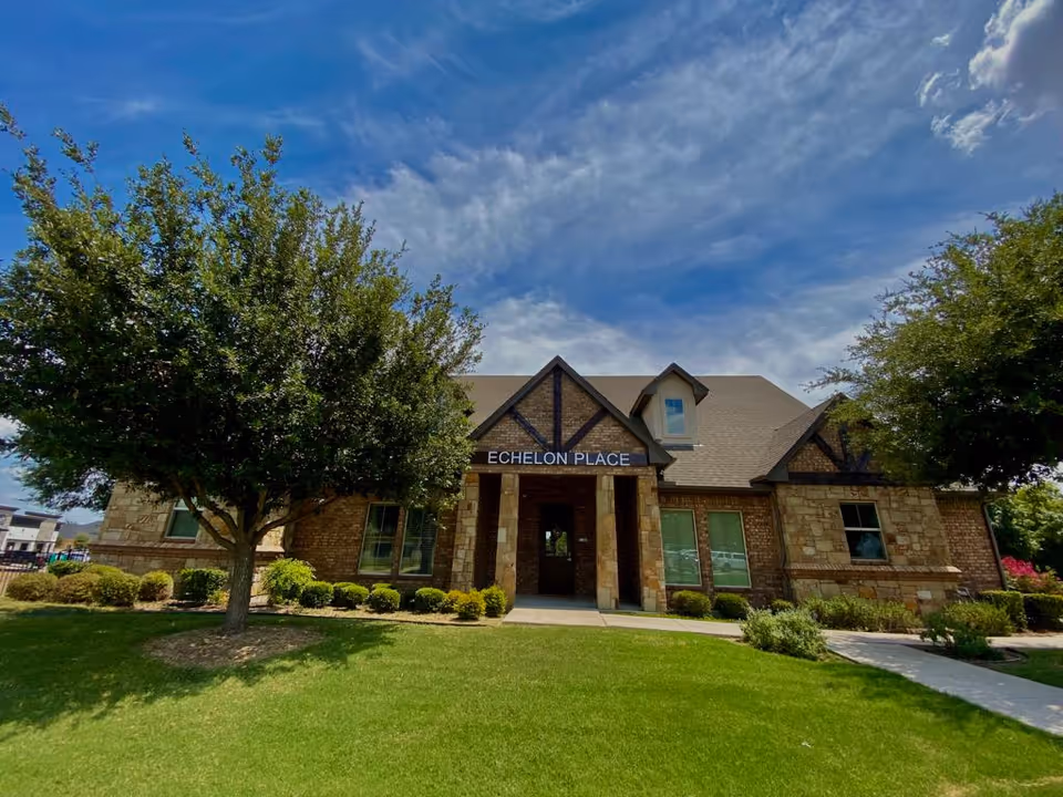 Front exterior view of Echelon Place Assisted Living building with a well-maintained lawn, trees, and shrubs under a partly cloudy blue sky.