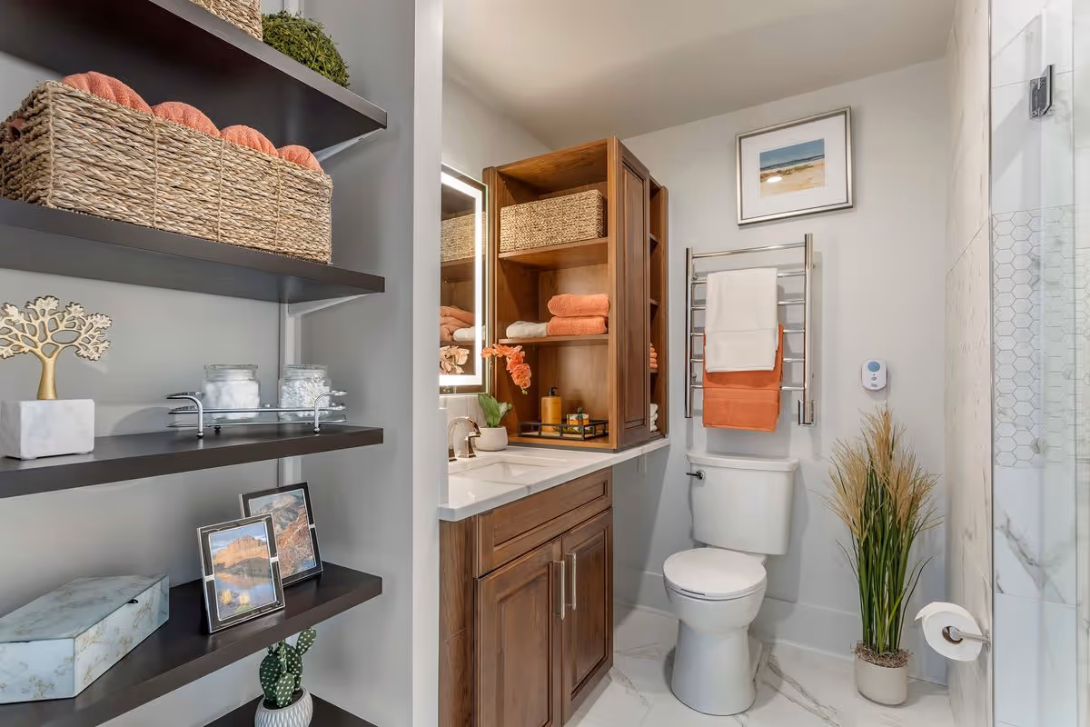 A modern bathroom featuring a white toilet with towels hanging on a metal rack above it, a wooden vanity with a white countertop and a mirror with built-in lighting, open wooden shelves holding folded towels and a basket, a glass-enclosed shower with hexagonal tile accents, and decorative items including framed pictures, plants, and storage baskets on dark floating shelves.