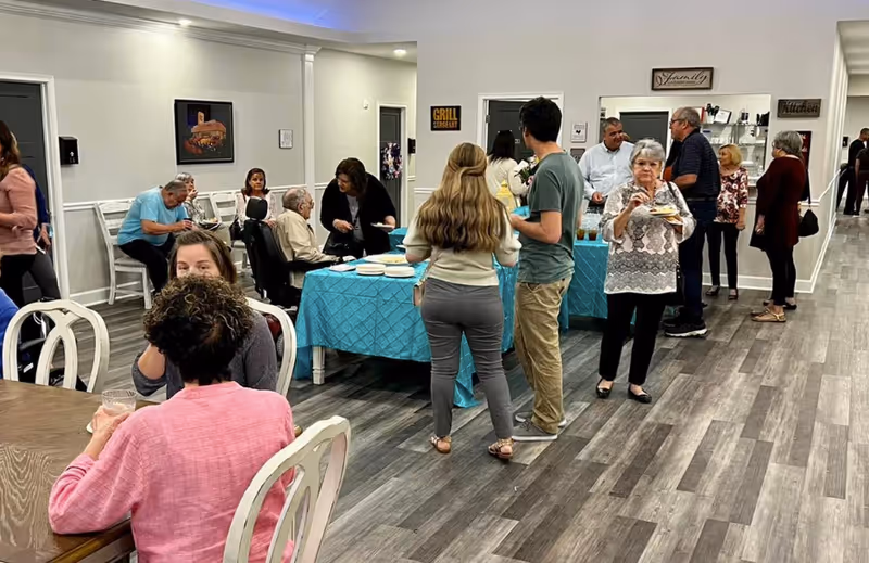 A group of people socializing and eating in a well-lit indoor space with gray wood flooring and white walls. There are tables with blue tablecloths holding food and plates. Some people are seated while others stand and chat near a serving area labeled 'Grill Sergeant' and 'Kitchen'.