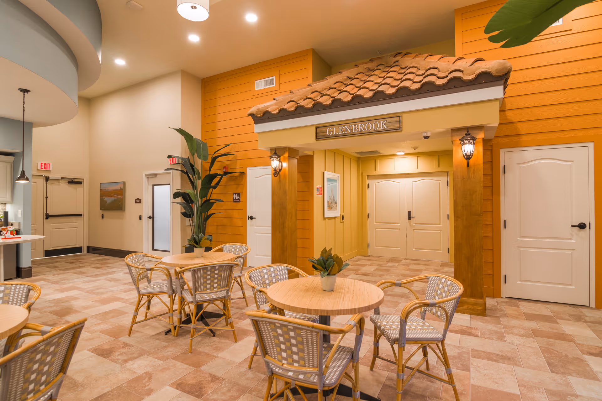 Interior view of a common area in a senior living facility with round wooden tables and wicker chairs arranged on a tiled floor. The walls are painted in warm tones with a section designed to look like a small house facade with a tiled roof and a sign that reads 'GLENBROOK'. There are potted plants on the tables and a large plant near the wall. Several doors and exit signs are visible in the background.