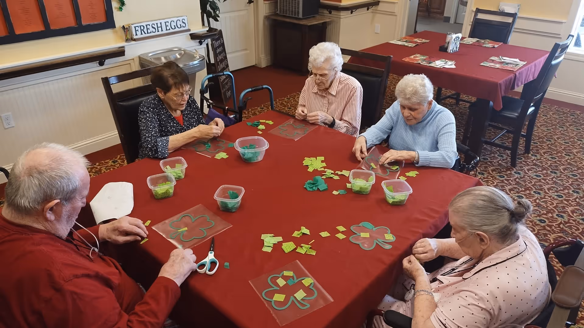 Five elderly individuals seated around a table covered with a red tablecloth, engaging in a craft activity involving green paper pieces and shamrock-shaped templates. The room has carpeted floors, additional tables with red tablecloths, and a sign on the wall that reads 'FRESH EGGS'.