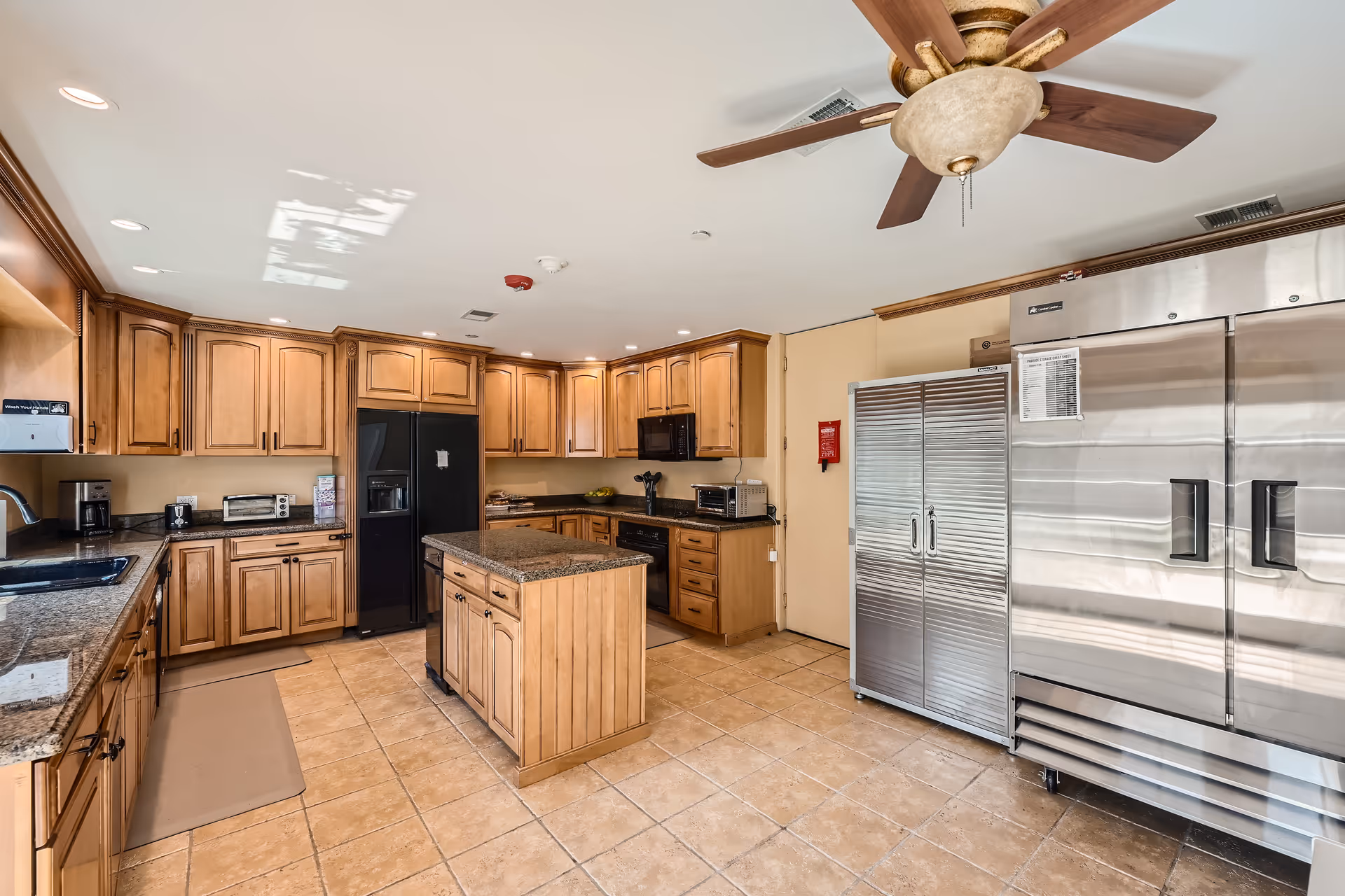 A spacious kitchen with wooden cabinetry, a central island, a black refrigerator, tile floor, and large stainless-steel commercial refrigerators.