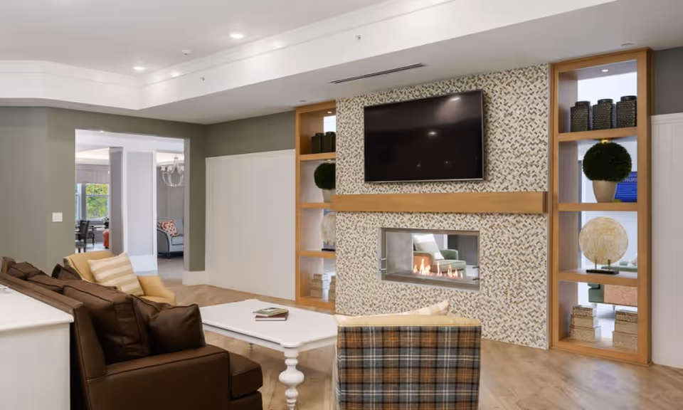 A cozy living room area featuring a brown leather sofa with striped pillows, a plaid armchair, and a white coffee table. The room has a modern fireplace set in a decorative tiled wall with built-in wooden shelves on either side holding various decorative items. Above the fireplace is a large flat-screen TV. The room has light wood flooring and an open doorway leading to another seating area.