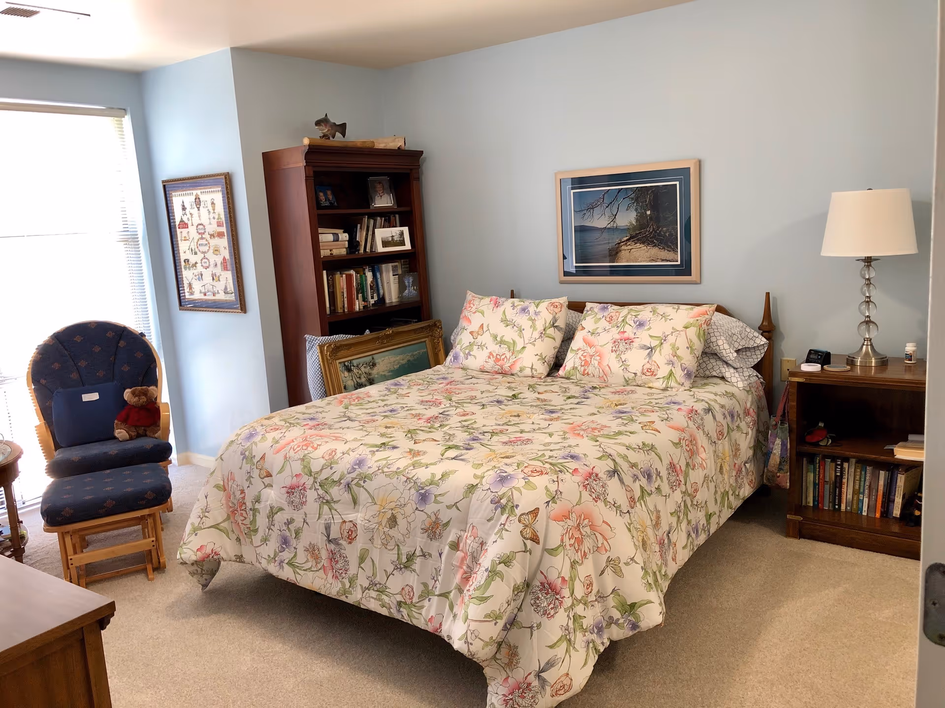 A cozy bedroom with light blue walls featuring a bed with floral patterned bedding and two matching pillows. To the right of the bed is a wooden nightstand with a lamp, books, and a few small items. Behind the bed is a framed picture of a beach scene. To the left of the bed is a tall wooden bookshelf filled with books and decorative items, with a framed picture leaning against it. Near the window is a cushioned rocking chair with a teddy bear sitting on it. The room is carpeted in a light beige color and has a window with blinds partially open.