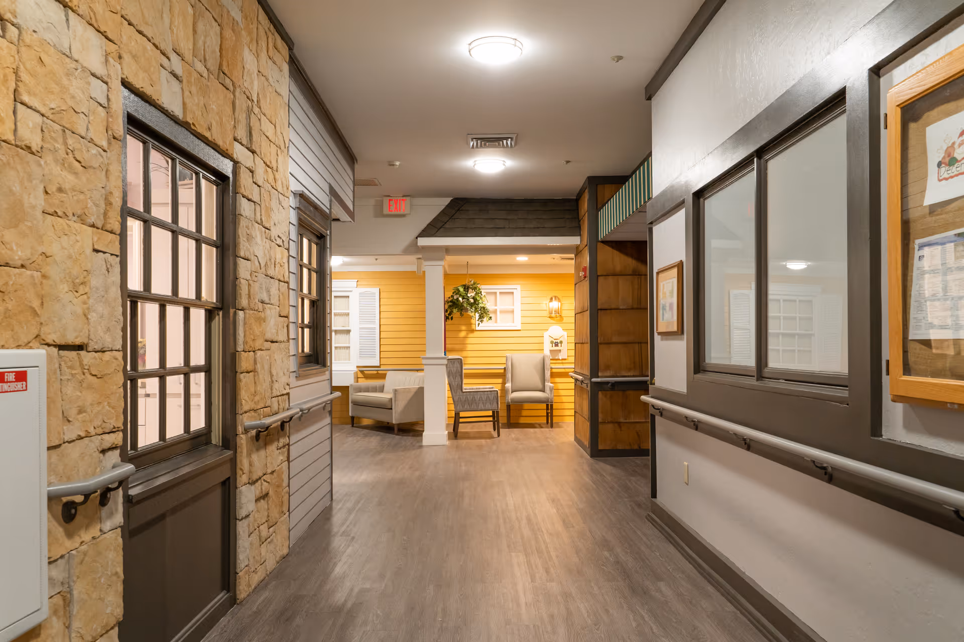 Indoor hallway in a senior living facility with stone and wood panel walls, handrails on both sides, and a seating area with chairs and a hanging plant at the end. The hallway is well-lit with ceiling lights and has an exit sign visible in the background.