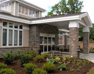 Exterior view of The Guilford House senior living facility showing a covered entrance with stone pillars, large windows, and landscaped garden beds with shrubs and small plants.