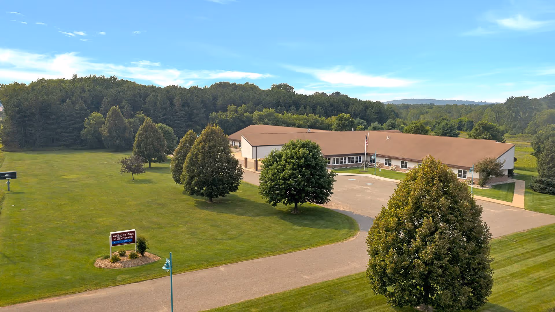 Aerial view of Wellington Place At Rib Mountain, a single-story building with a brown roof surrounded by green lawns and trees under a blue sky with some clouds.