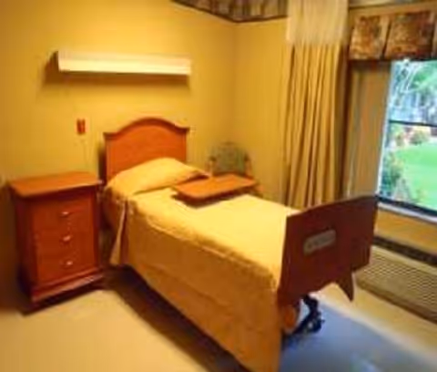 A small, simple bedroom in a rehabilitation center featuring a single bed with a wooden headboard and footboard, a beige bedspread, a wooden bedside table with drawers, a chair in the corner, beige curtains covering a window, and a wall-mounted light fixture above the bed.