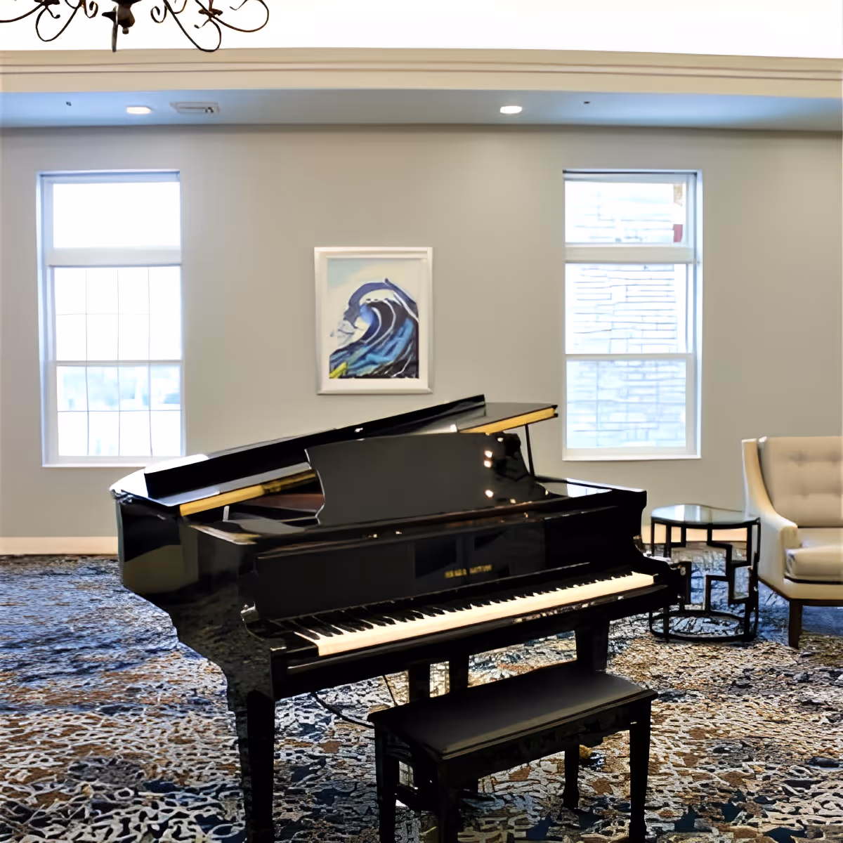 A black grand piano with a matching bench is placed on a patterned carpet in a well-lit room. The room has two large windows with white blinds, a framed painting of a wave on the wall, a beige armchair, and a small round glass-top side table.