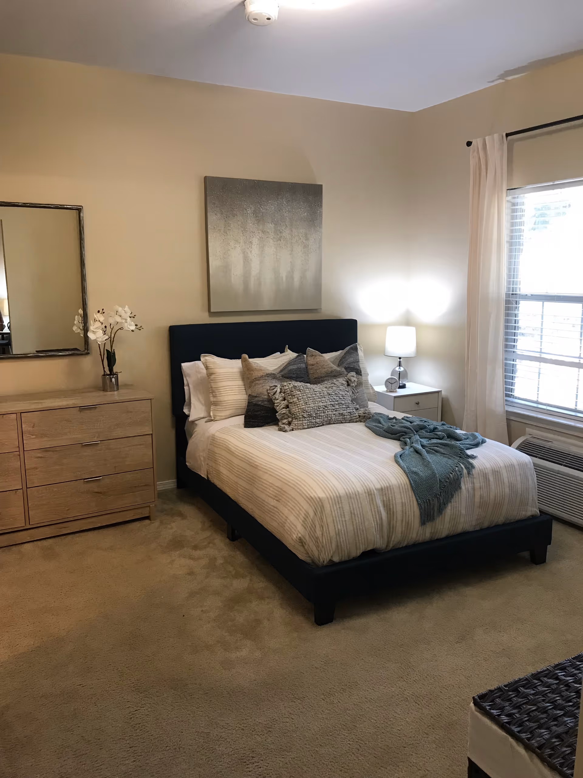 Neatly made bedroom with a bed, decorative pillows and throw, bedside table with lamp, dresser and window with blinds.