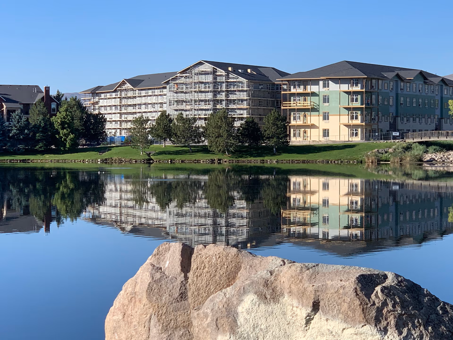 Multi-story residential buildings (one with scaffolding) reflected in a calm lake with large rocks in the foreground.