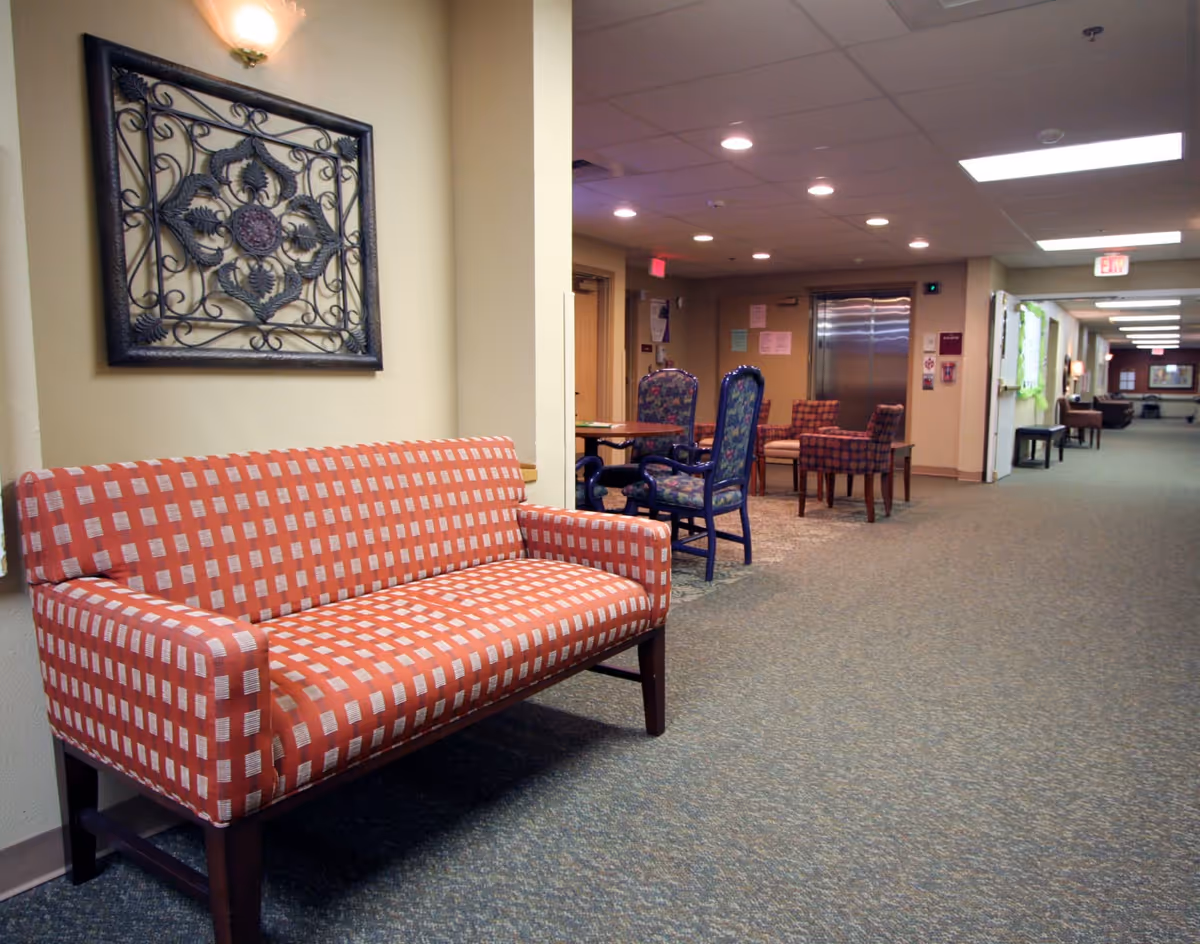 Carpeted hallway lounge in a senior living facility with a patterned bench in the foreground and tables, chairs and an elevator in the background.