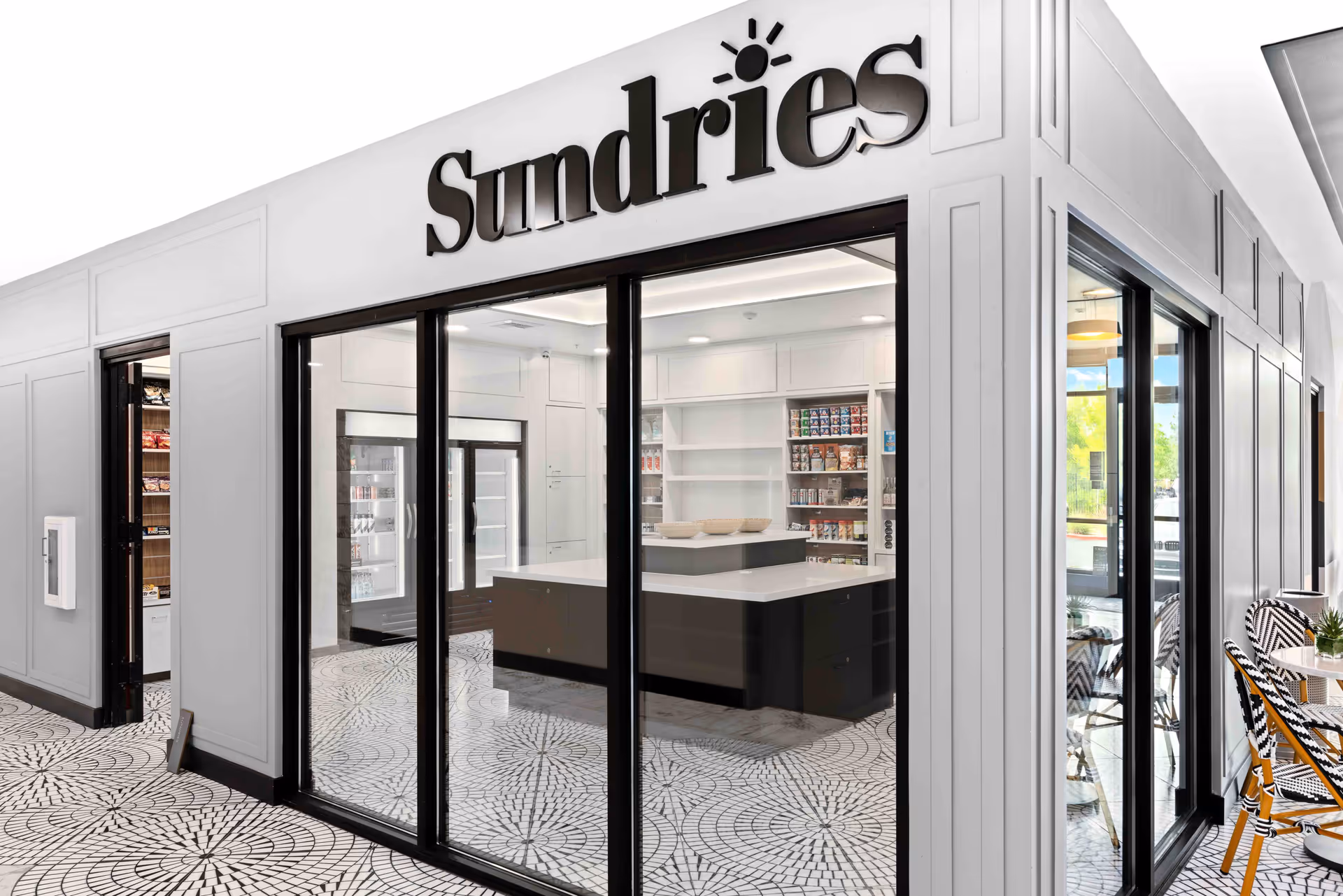 Interior view of a sundries shop area in a senior living facility named Revel Folsom, featuring glass doors and windows, a central counter, shelves stocked with various items, and a patterned tile floor.