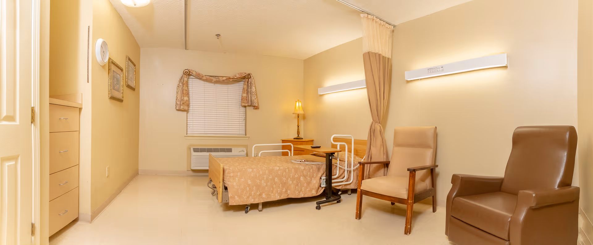 A nursing facility room with a single bed covered in a beige patterned blanket, two chairs including one brown leather and one wooden with beige cushions, a small bedside table with a lamp, a window with blinds and a decorative valance, and a privacy curtain partially drawn. The walls and floor are light-colored, and there is an air conditioning unit below the window.