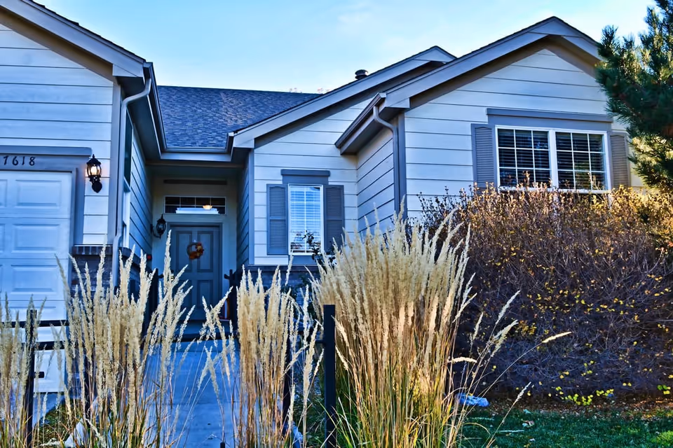 Exterior view of a single-story residential building with light gray siding, a front door with a wreath, windows with shutters, and ornamental grasses and bushes in the front yard under a clear blue sky.