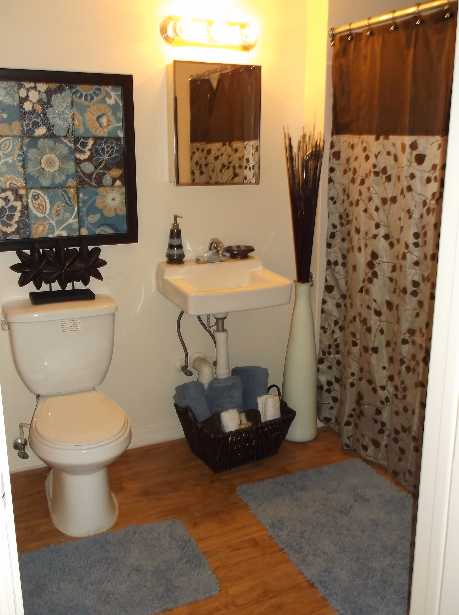 A bathroom with a white toilet and a small white wall-mounted sink. Above the sink is a rectangular mirror with a light fixture above it. To the right is a shower with a brown and beige patterned curtain. On the floor are two blue bath mats, and a basket with rolled towels is placed under the sink. A tall vase with decorative sticks is next to the shower. A framed floral artwork hangs on the wall above the toilet.