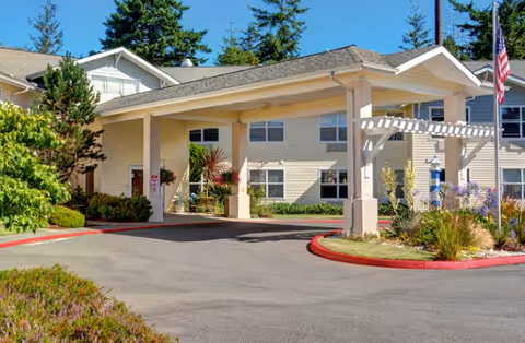 Entrance of a senior living facility with a covered porte-cochere, driveway, landscaping, and an American flag.