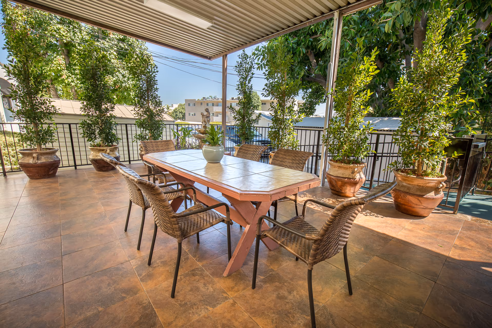 Outdoor covered patio area with a rectangular table surrounded by six wicker chairs. The patio is tiled and decorated with large potted plants along the railing, with trees and buildings visible in the background under a clear blue sky.