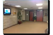 Carpeted interior lobby with a wall-mounted TV, potted plants, framed art, and double doors at the far end.