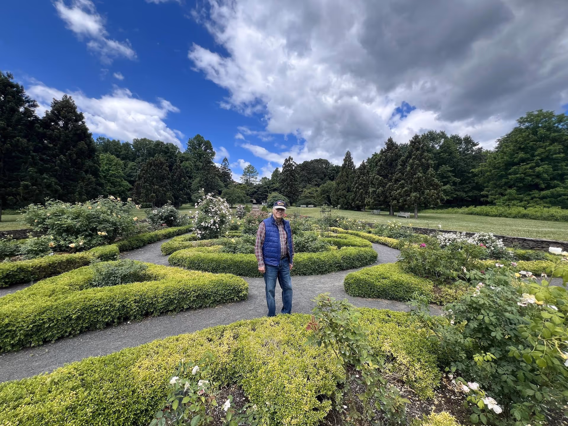 An elderly man wearing a blue vest, plaid shirt, jeans, and a cap stands in the middle of a manicured garden with neatly trimmed hedges and flowering bushes. The garden is surrounded by tall trees under a partly cloudy sky.
