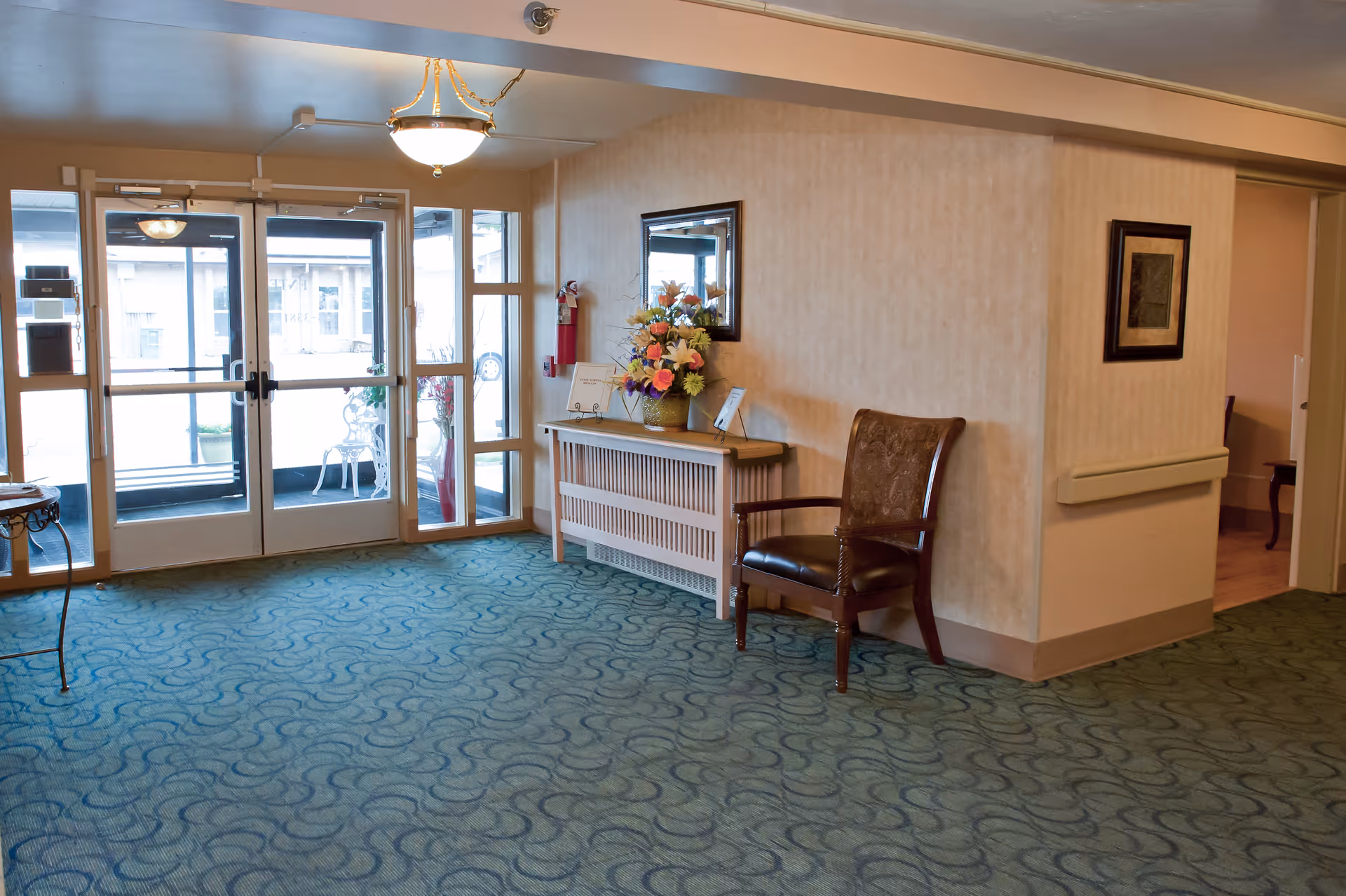 Interior view of a senior living facility entrance area with double glass doors, a green patterned carpet, a wooden chair with armrests, a console table with a flower arrangement, a mirror on the wall, and a fire extinguisher mounted on the wall.