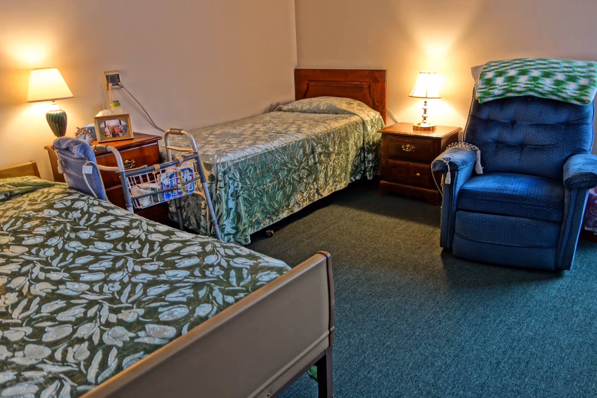 Two twin beds with green bedspreads, nightstands and lamps, a walker, and a blue recliner in a senior living bedroom.