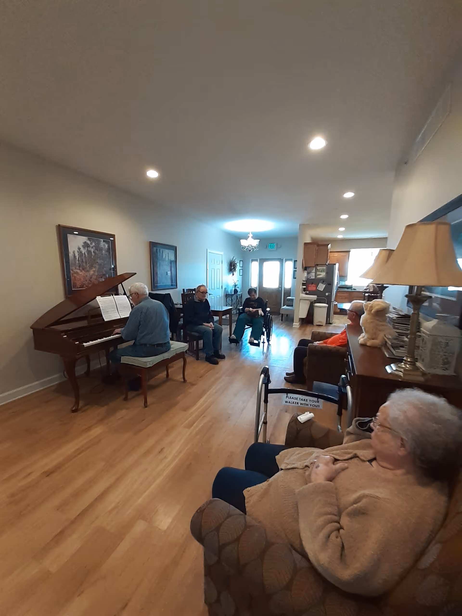A group of elderly people in a living room setting at Pinnacle Memory Care in Muscatine IA. One person is playing a grand piano while others are seated on chairs and couches, some using walkers. The room has wooden flooring, framed artwork on the walls, and a lamp on a side table.