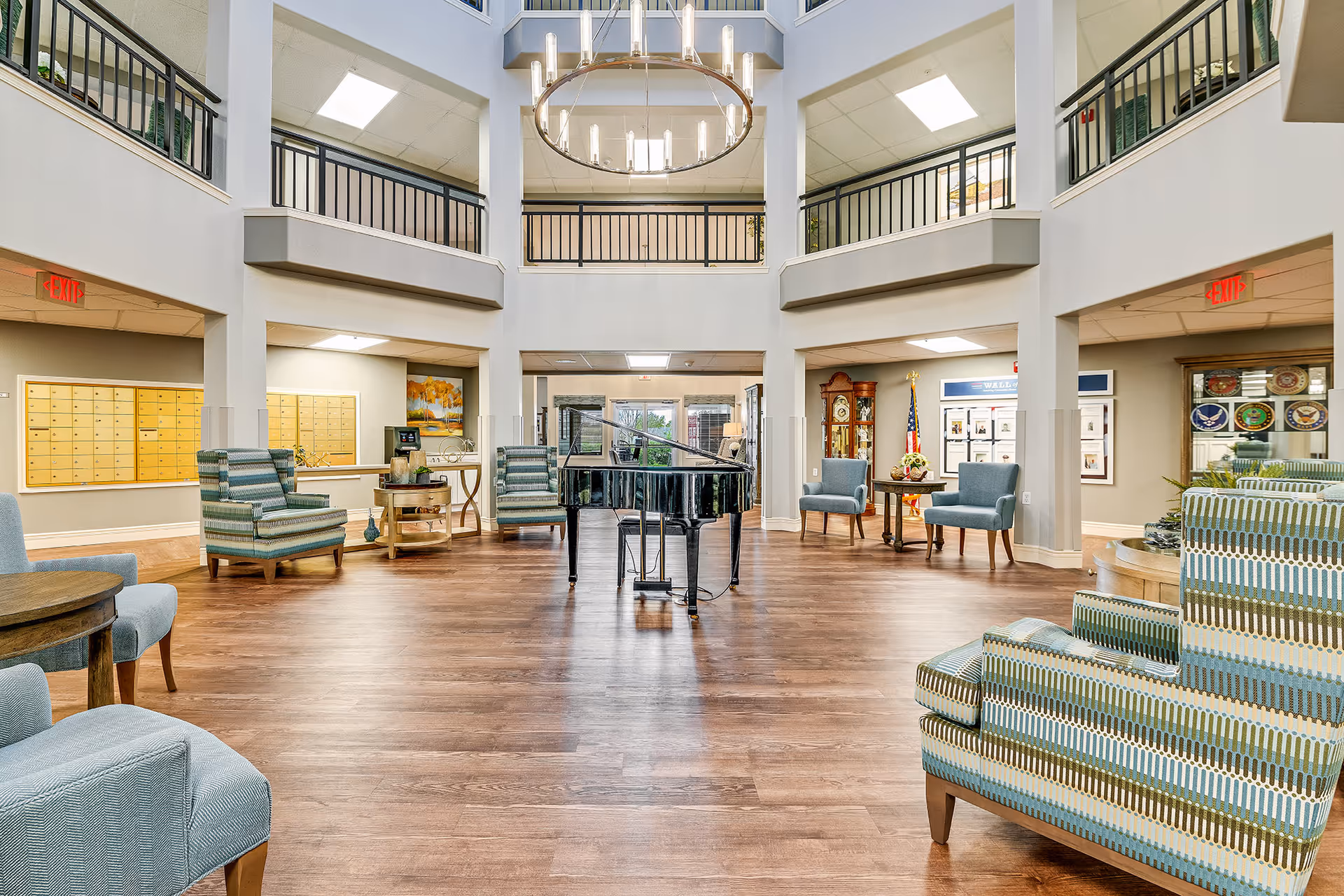 Spacious senior living facility common area with a grand piano in the center, surrounded by multiple upholstered chairs and small tables. The room features high ceilings with a large circular chandelier, wooden flooring, and a second-floor balcony with black railings. There are exit signs, a grandfather clock, an American flag, and a wall with framed pictures in the background.