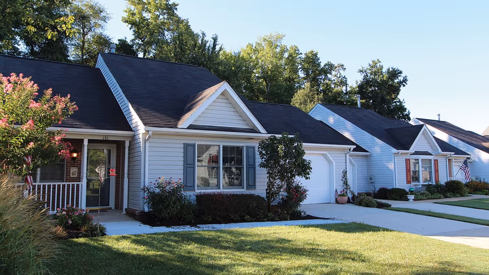 Exterior view of single-story residential homes with well-maintained lawns, shrubs, and trees under a clear blue sky.
