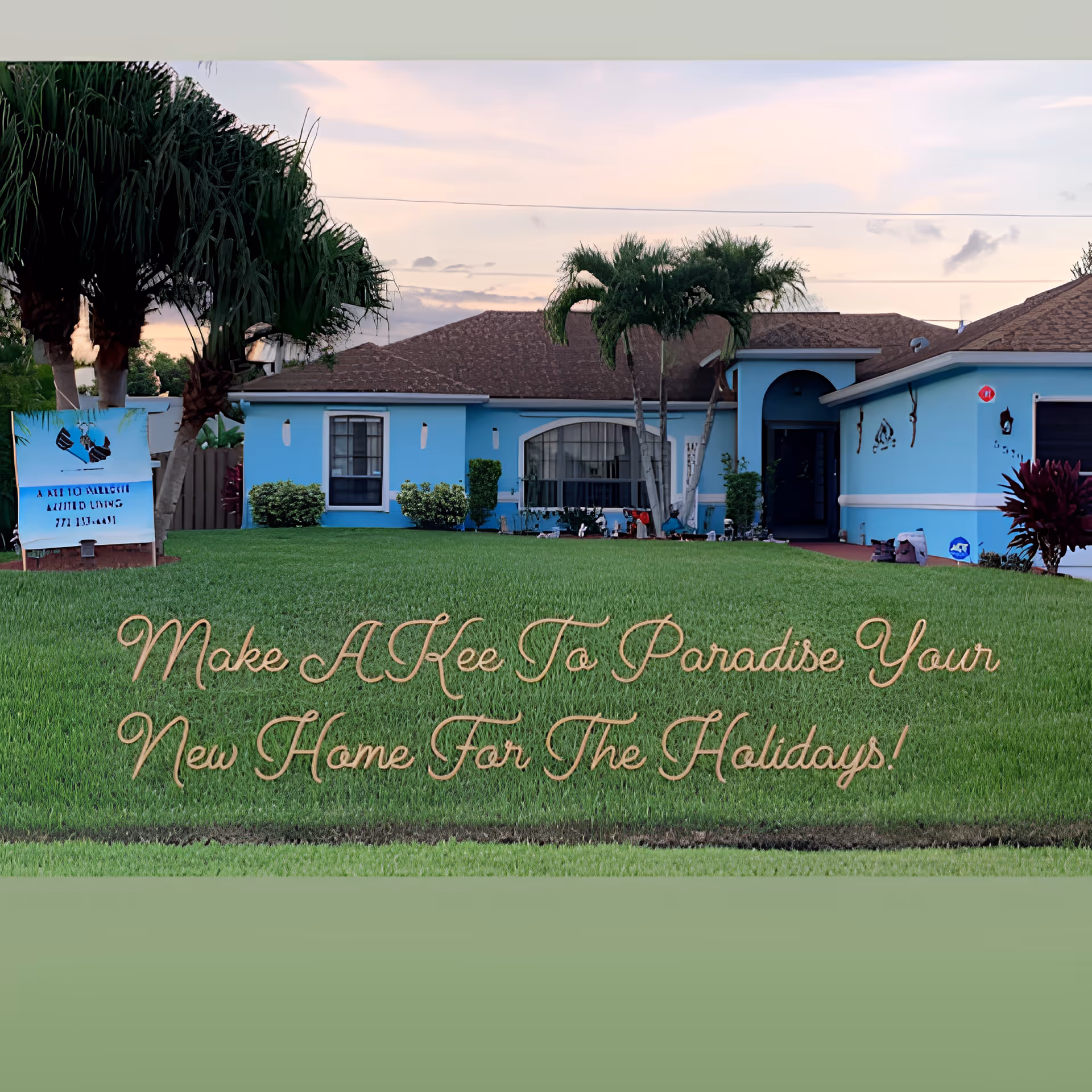 A single-story blue house with a brown roof surrounded by a well-maintained green lawn and palm trees under a partly cloudy sky at sunset. There is a sign on the left side of the lawn with text and images, and decorative items near the house entrance. Text on the lawn reads, 'Make A Kee To Paradise Your New Home For The Holidays!'