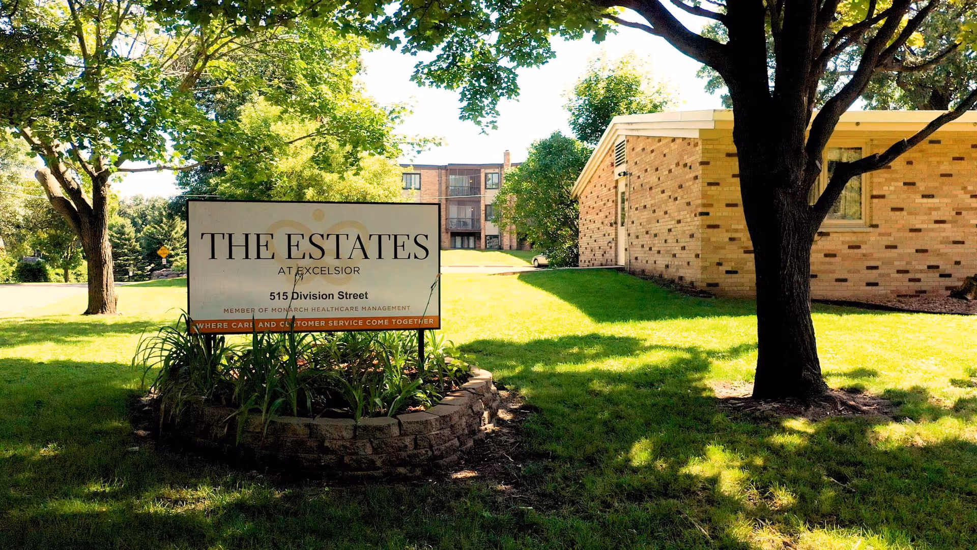 Outdoor view of The Estates at Excelsior sign on a grassy lawn with trees and a brick building in the background.
