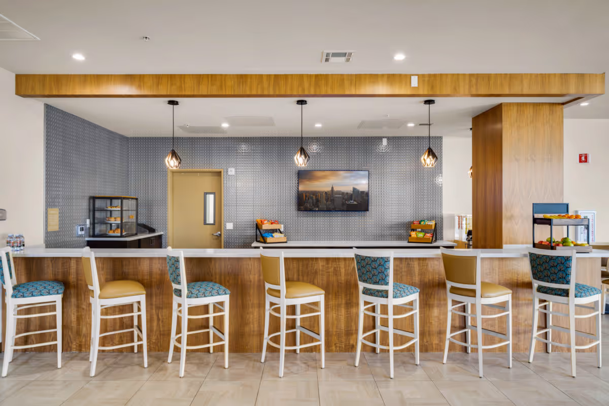 A modern snack bar area with a long wooden counter and seven high chairs, some with blue patterned cushions and others with tan cushions. Behind the counter is a gray textured wall with a mounted flat-screen TV displaying a cityscape. There are three pendant lights hanging from the ceiling, and various snacks and fruits are displayed on the counter and shelves.