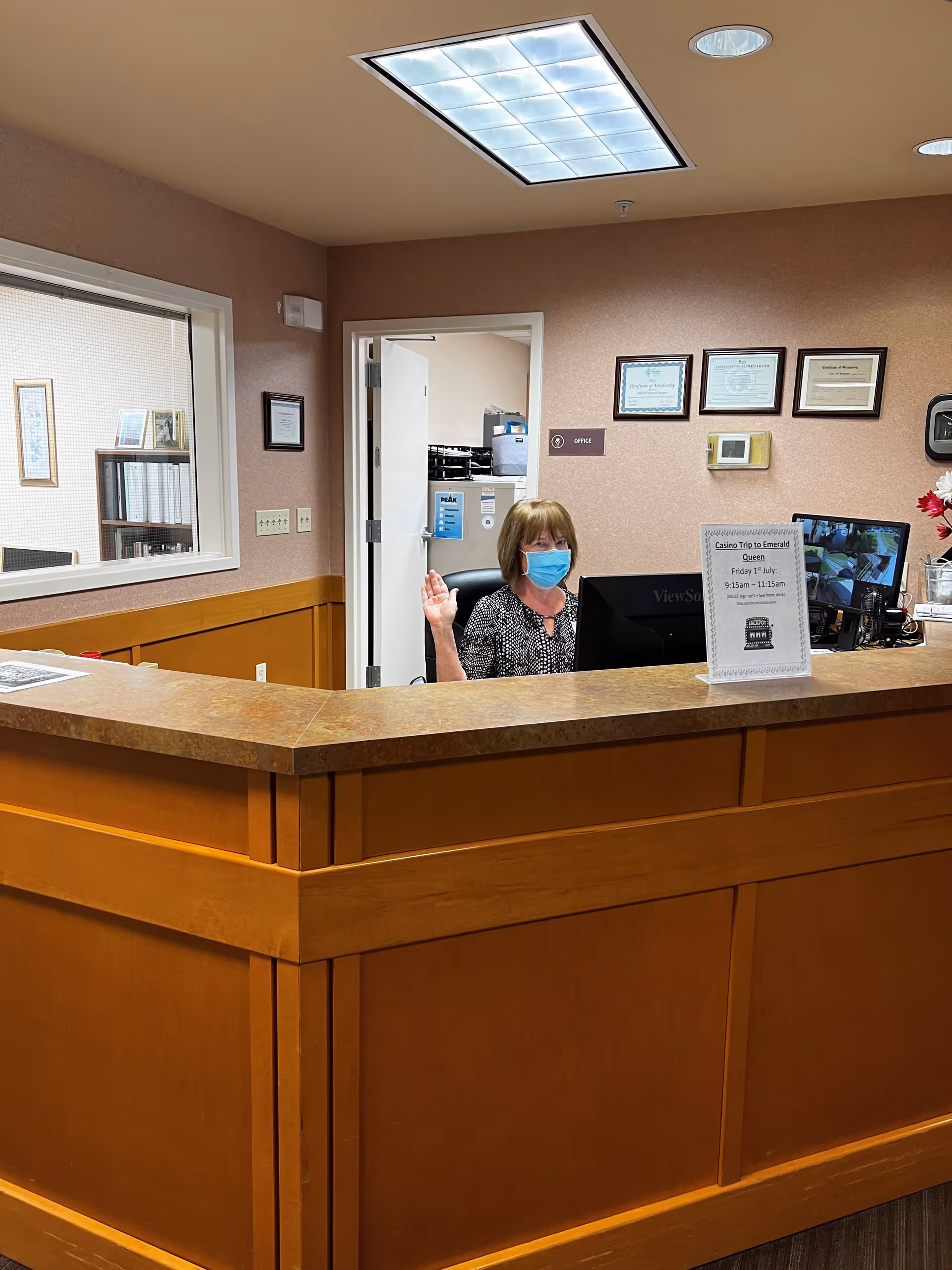 Reception area with a wooden front desk and a woman wearing a face mask sitting behind the desk waving. The wall behind her has framed certificates and a sign labeled 'Office'. There is a window to another room on the left side and a ceiling light above.