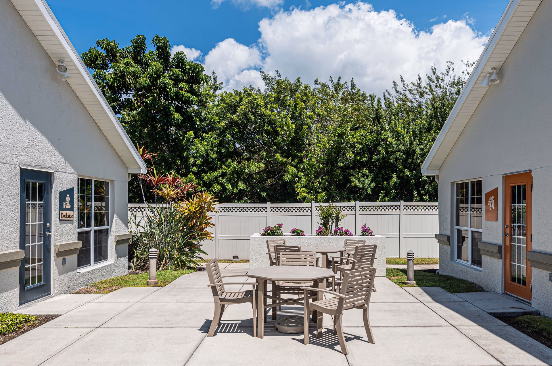 Outdoor patio area between two white buildings with a round table and six chairs on a concrete surface, surrounded by greenery and a white fence under a blue sky with clouds.