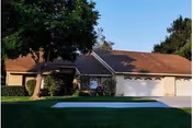 Single-story residential building with a brown shingle roof, beige exterior walls, and a three-car garage. The front yard features a well-maintained green lawn, a large tree on the left side, and some shrubs near the house. The sky is clear and blue.