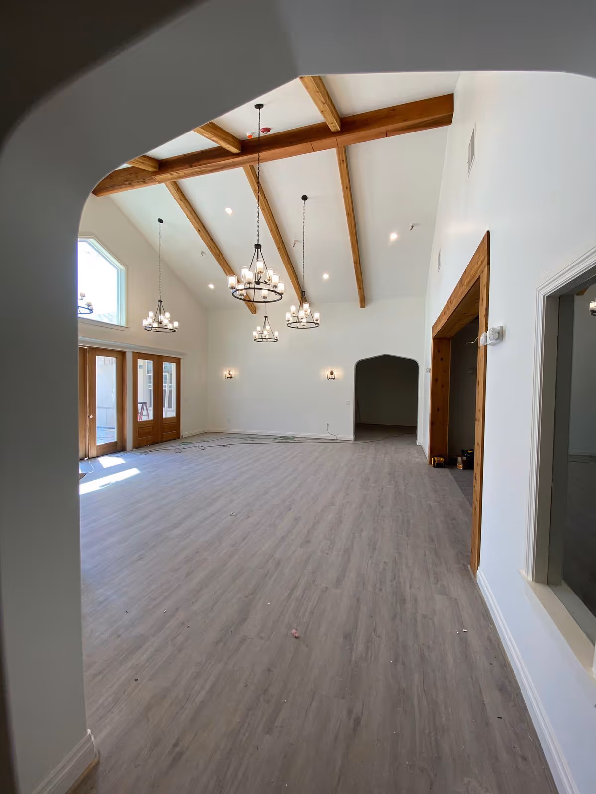 A spacious interior room with high vaulted ceilings featuring exposed wooden beams and multiple hanging chandeliers. The room has light-colored wood flooring, white walls, and large windows and glass doors letting in natural light. There is an arched doorway at the far end and a large open doorway with wooden trim on the right side.