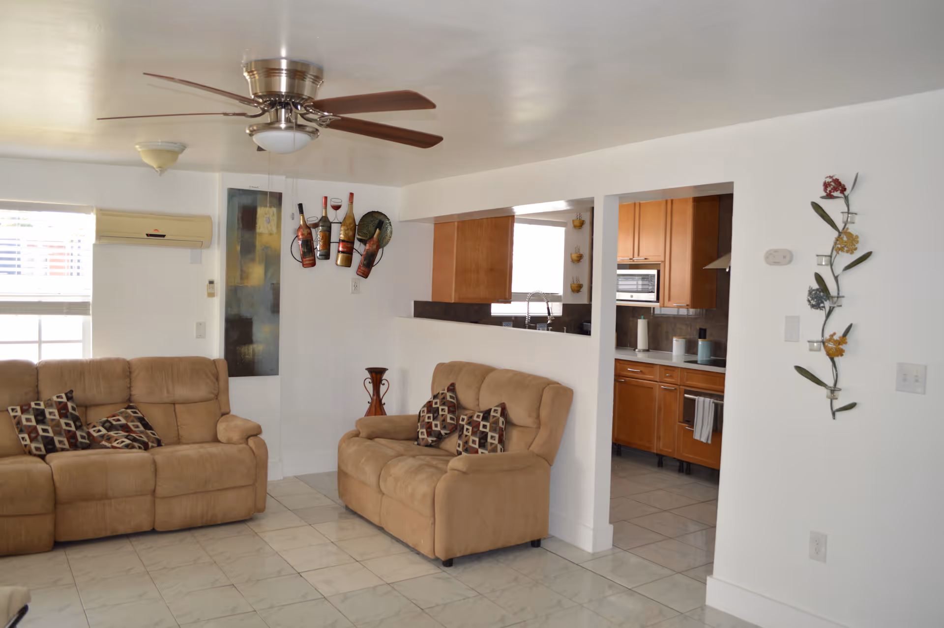 A cozy living room with two beige recliner sofas adorned with patterned cushions. The room has white walls and a tiled floor. A ceiling fan with wooden blades is mounted on the ceiling. There is a window with blinds and an air conditioning unit on the wall. The living room opens into a kitchen area with wooden cabinets and a countertop visible through a large pass-through window. Decorative wall art and a tall vase are also present.