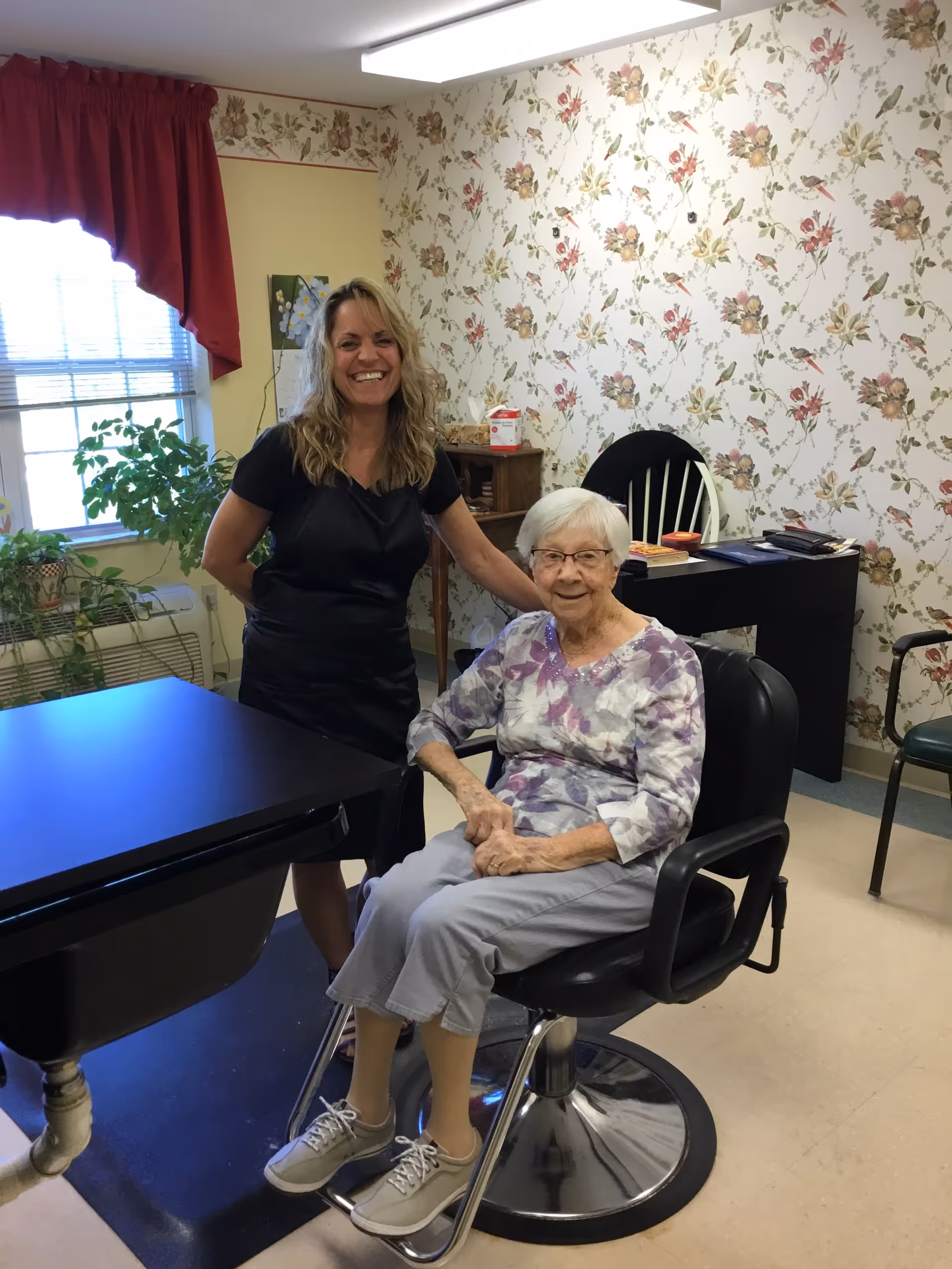 An elderly woman with white hair and glasses sits in a black salon chair smiling, wearing a purple and white patterned top and light gray pants. A woman with long blonde hair, dressed in black, stands behind her smiling. The room has floral wallpaper, a window with red curtains, a black table, and some plants near the window.