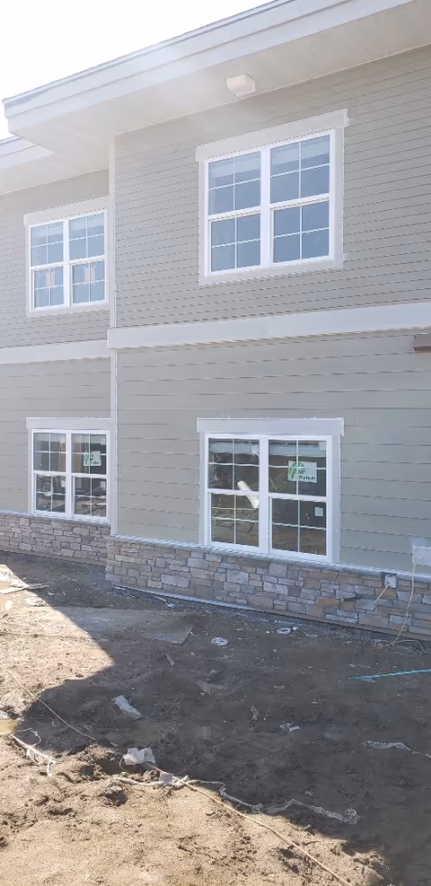 Exterior facade of a senior living building under construction with white-framed windows, gray horizontal siding, stone veneer at the base, and bare dirt in front.