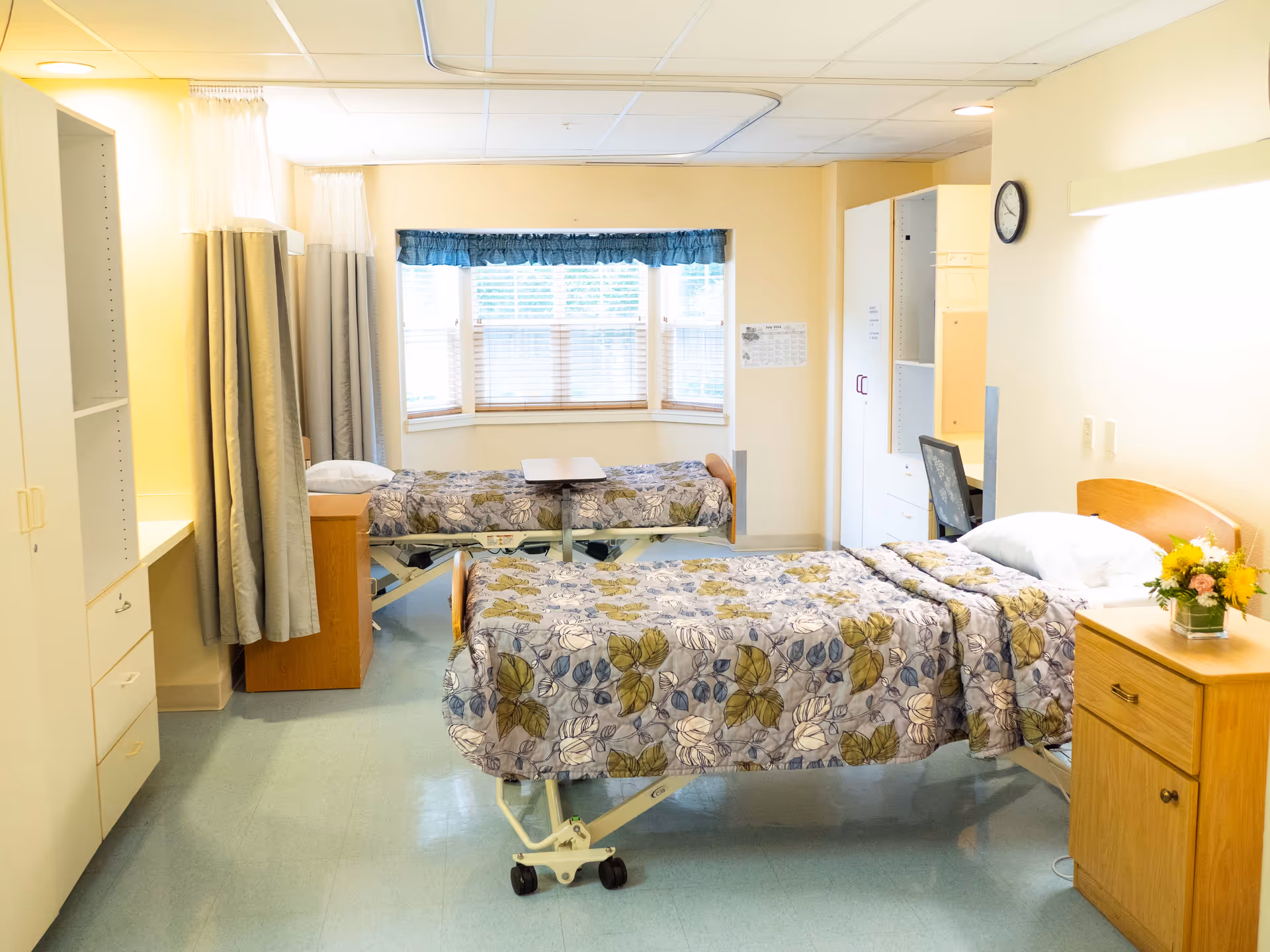 A bright and clean room with two hospital-style beds covered with floral-patterned bedspreads. The room has a large window with blinds and a blue valance, a wooden nightstand with a flower arrangement, a clock on the wall, and a desk with a chair. There are curtains between the beds for privacy and built-in storage cabinets along one wall.