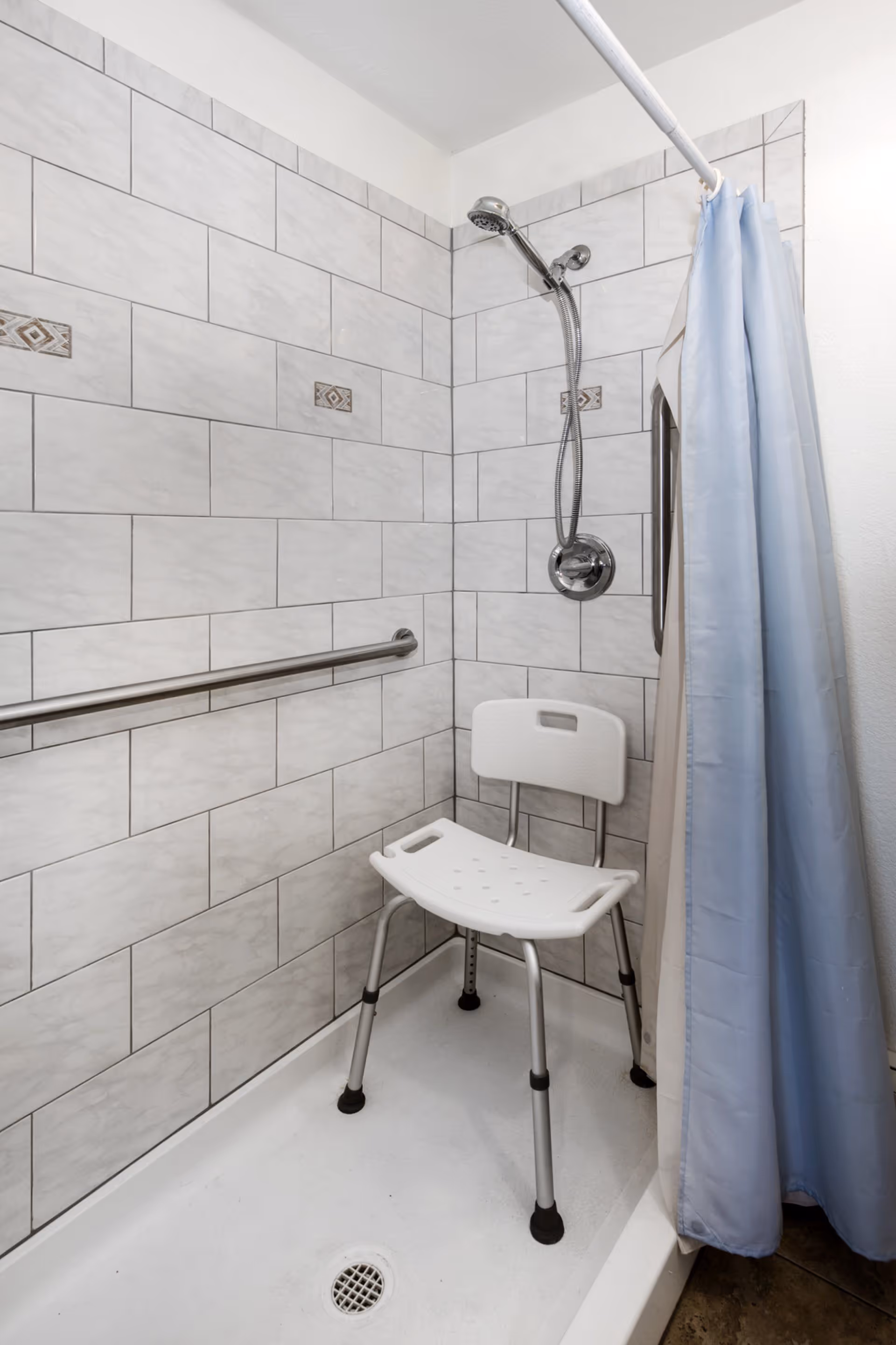 A shower area with white tiled walls featuring decorative accent tiles, a silver showerhead with a flexible hose, a white shower chair with a backrest, a horizontal grab bar on the wall, and a light blue shower curtain partially drawn to the side.